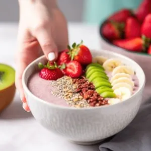 Smoothie bowl topped with sliced banana, kiwi, strawberries, and flaxseed, with a hand reaching in to garnish.