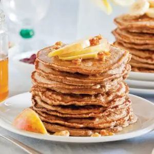 Stack of golden-brown organic multigrain pancakes topped with chopped walnuts and citrus slices, served on a white plate with a peach wedge.